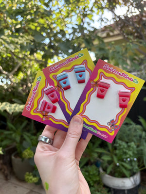 Hand holding three pairs of coffee cup polymer clay earrings on display cards with a blurred outdoor background
