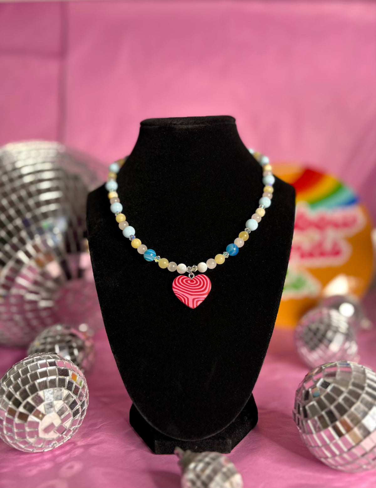 A beaded necklace with pastel blue, yellow, and white beads and a pink striped heart charm, displayed on a black velvet bust against a pink background with disco ball props.