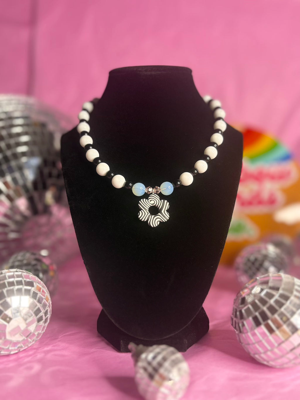 A beaded necklace displayed on a black velvet necklace bust against a pink background, with disco ball props around it. The necklace features white round beads, two opal beads, and a central black and white pendant with black zebra-like stripes.