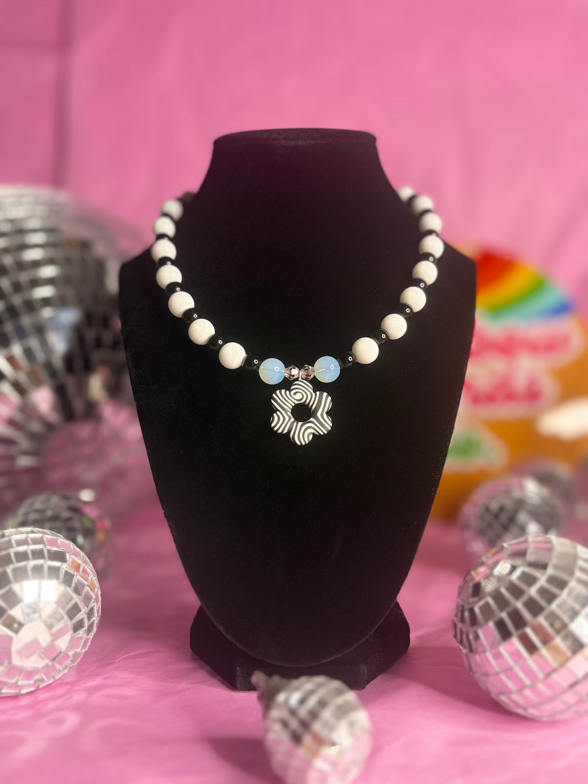 A beaded necklace displayed on a black velvet necklace bust against a pink background, with disco ball props around it. The necklace features white round beads, two opal beads, and a central black and white pendant with black zebra-like stripes.