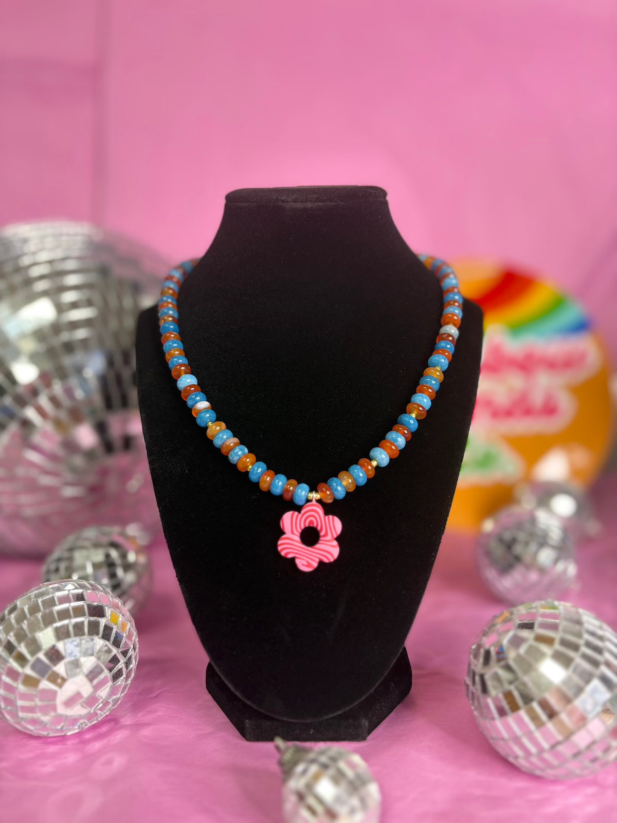 A beaded necklace displayed on a black velvet jewelry bust against a pink background, decorated with disco balls. The necklace features light blue and orange beads with a pink flower-shaped charm.
