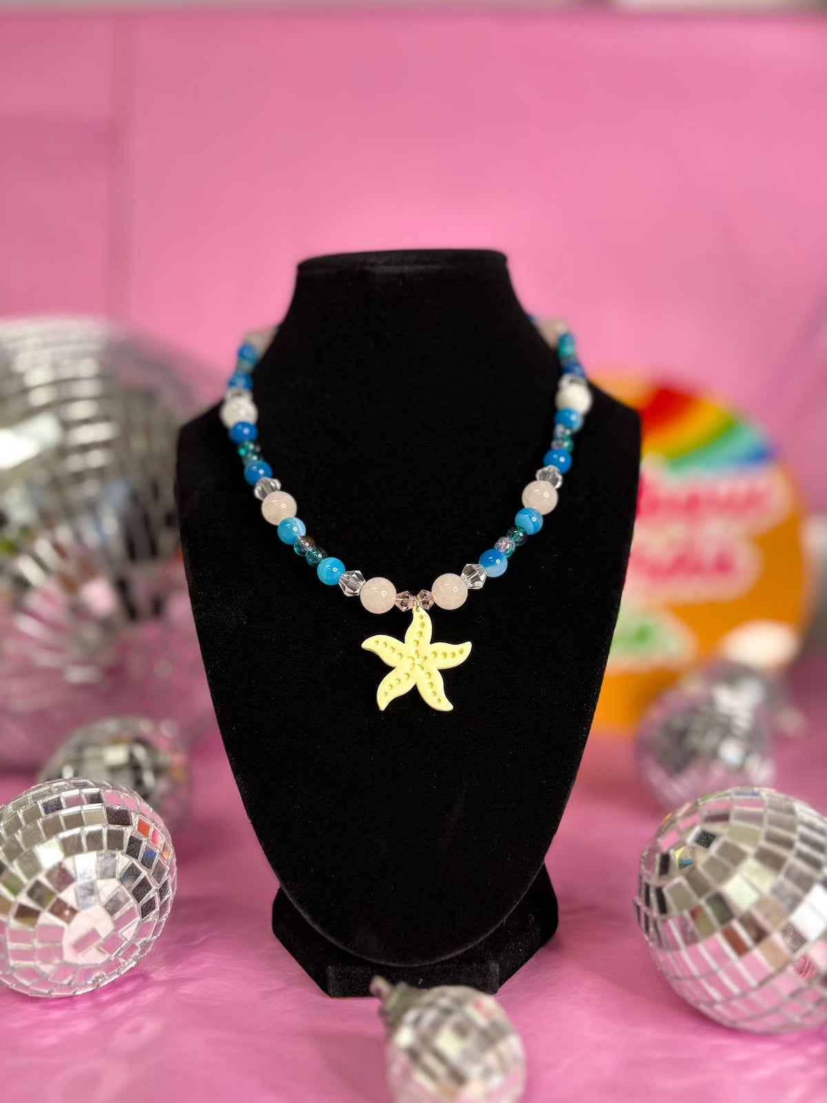 A beaded necklace with blue, white, and clear beads and a yellow starfish pendant, displayed on a black velvet necklace bust against a pink background with decorative disco balls.