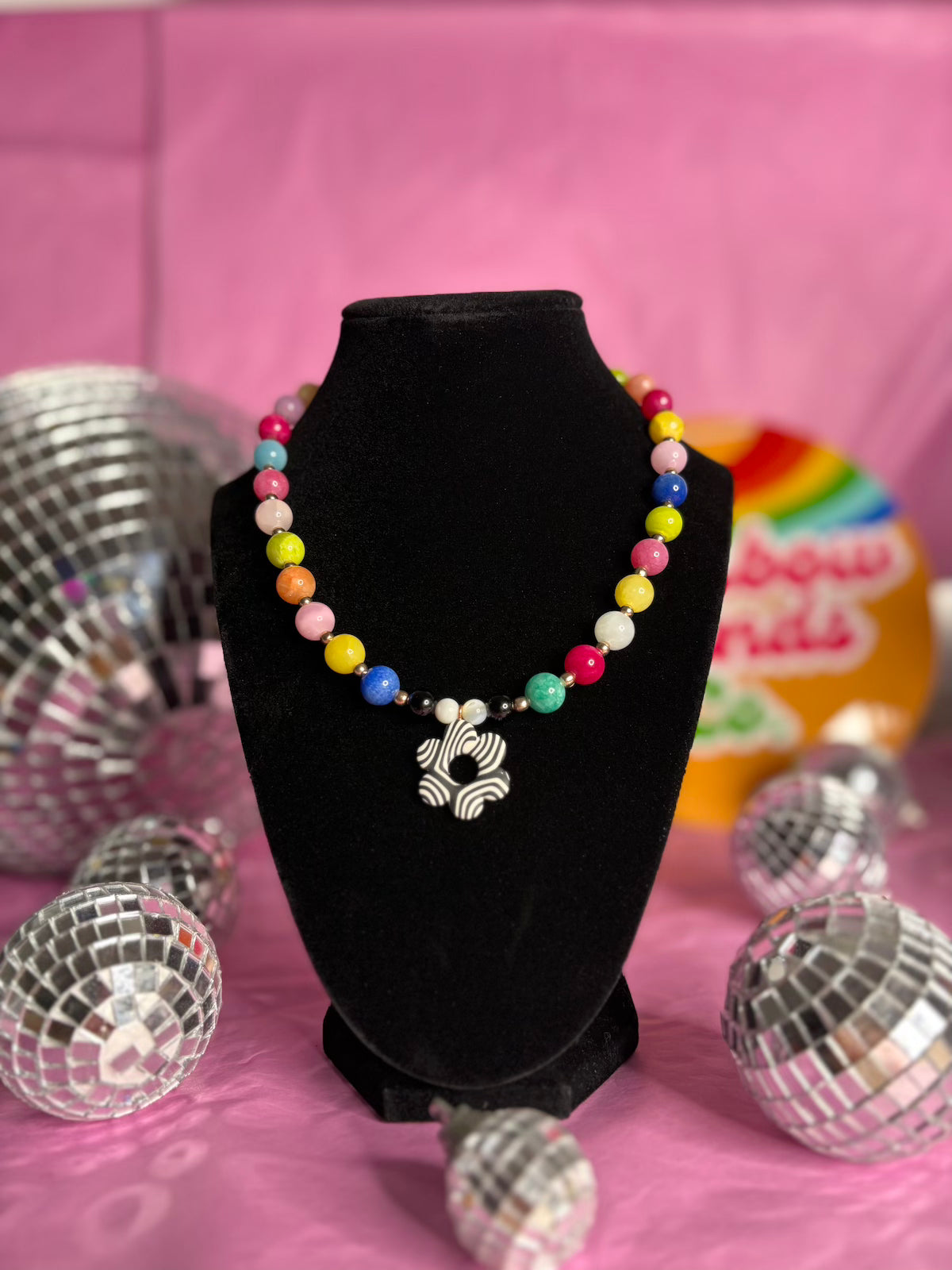 A colourful beaded necklace with a black and white flower polymer clay  pendant displayed on a black velvet necklace bust, surrounded by disco balls on a pink background.