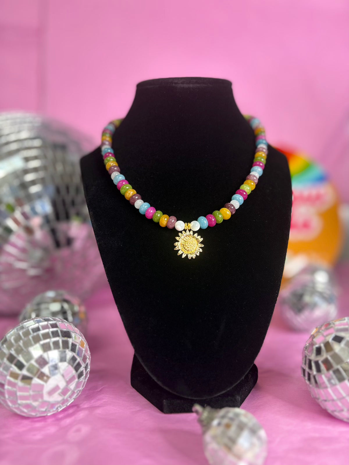 A colorful beaded necklace with a gold sun pendant displayed on a black velvet bust against a pink background with disco ball props.