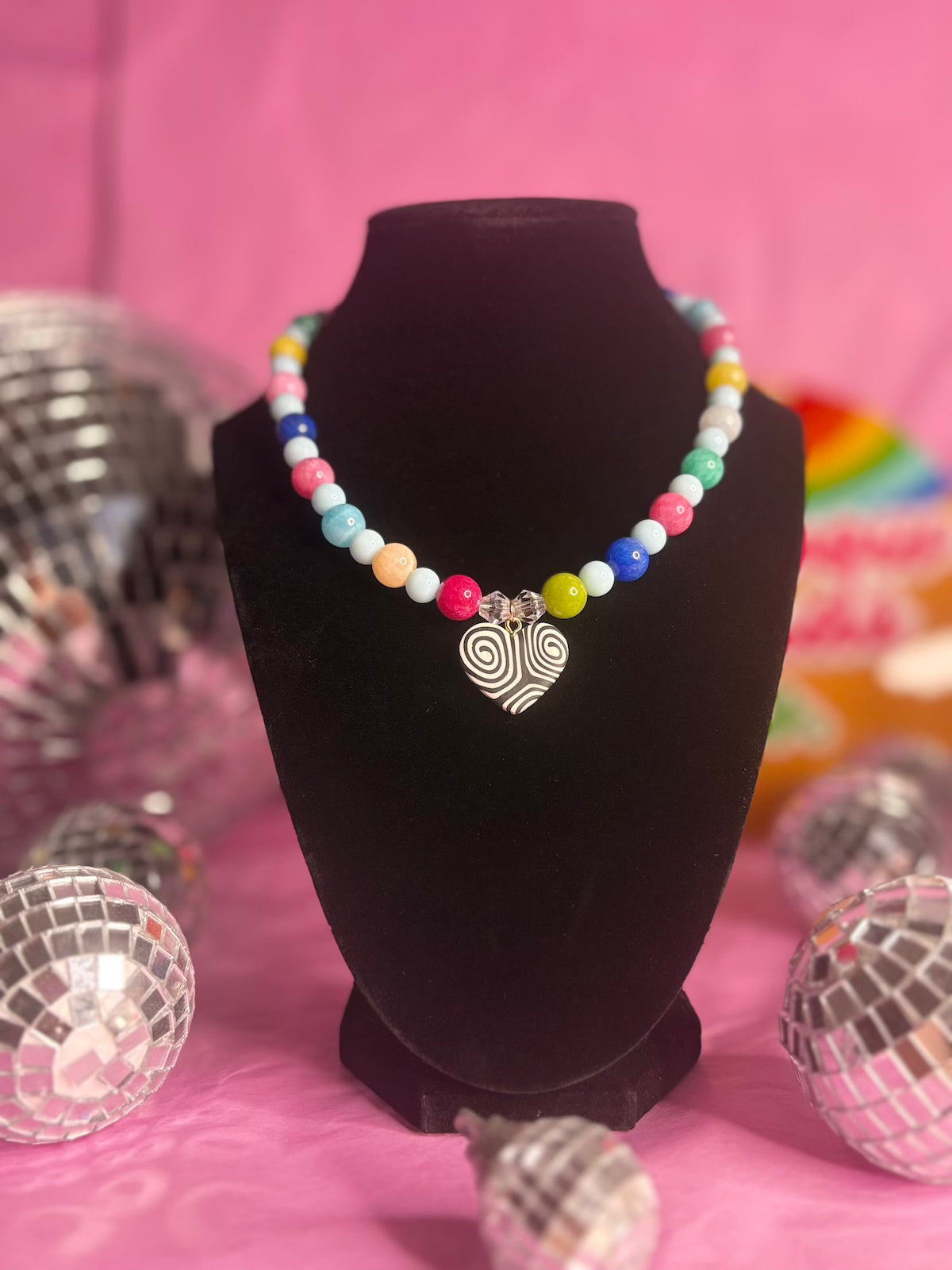 A colorful beaded necklace with a silver spiral heart pendant, displayed on a black velvet jewelry bust against a pink background with disco ball props.