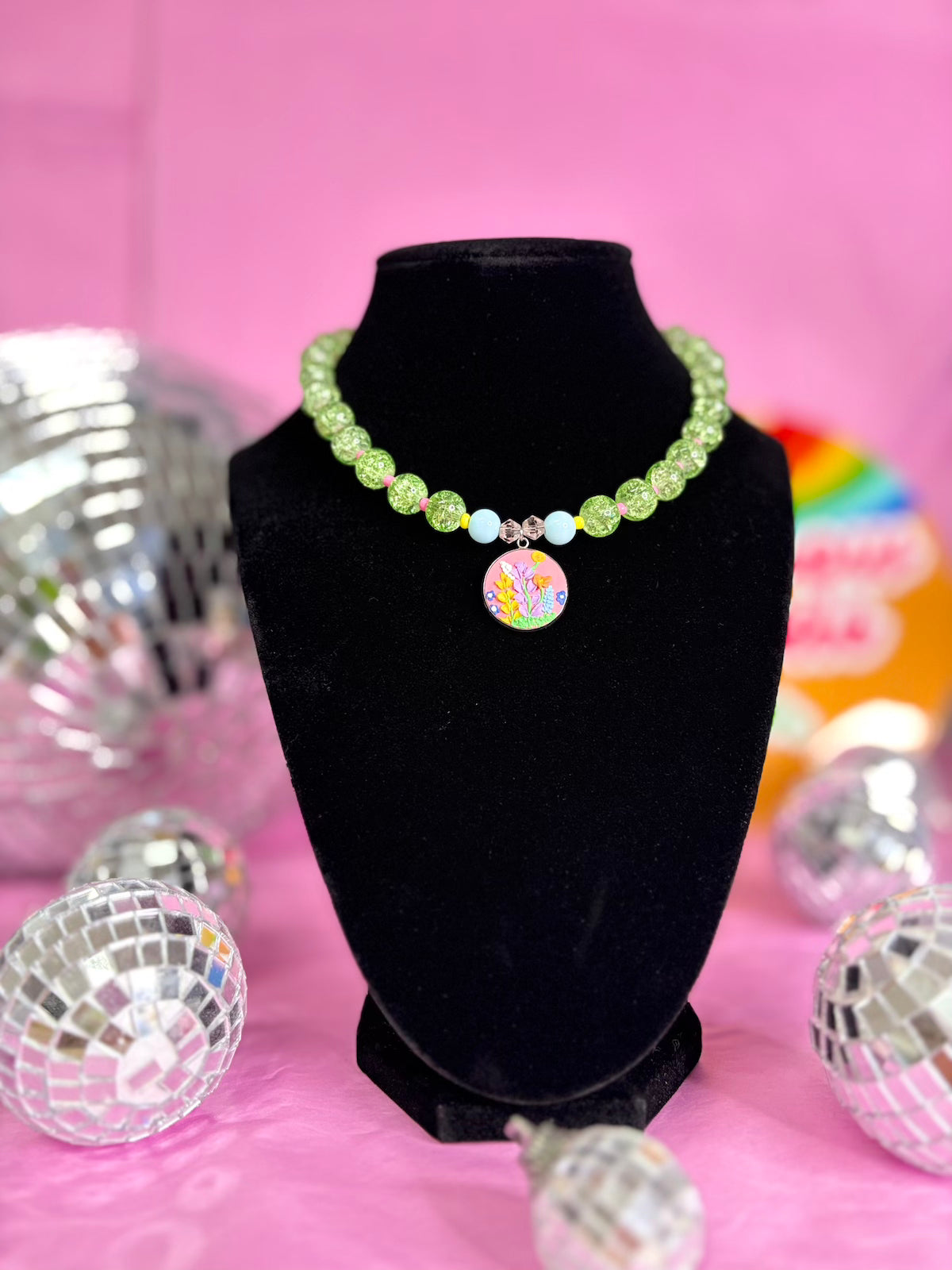 A beaded necklace with light green beads, small white and blue beads, and a colorful round pendant, displayed on a black velvet stand against a pink background with disco ball props.