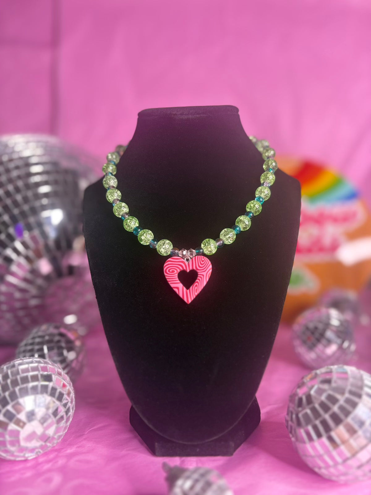 A beaded necklace with green faceted beads and a pink striped heart pendant, displayed on a black jewelry bust against a pink background with disco ball props.