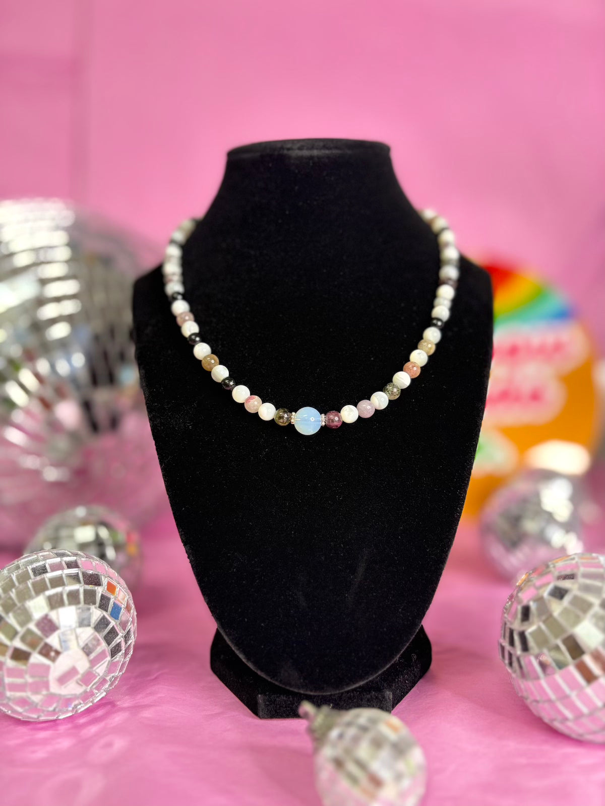 A beaded necklace displayed on a black velvet jewelry bust against a bright pink background, surrounded by mirrored disco balls.