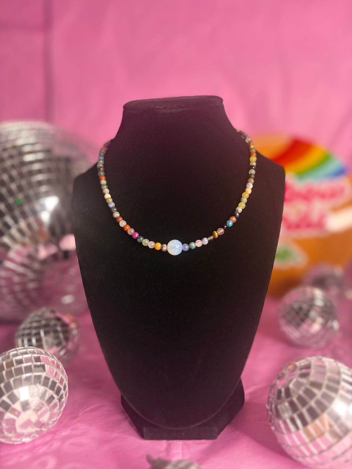 A colorful beaded necklace displayed on a black velvet jewelry bust against a pink background with disco ball props.