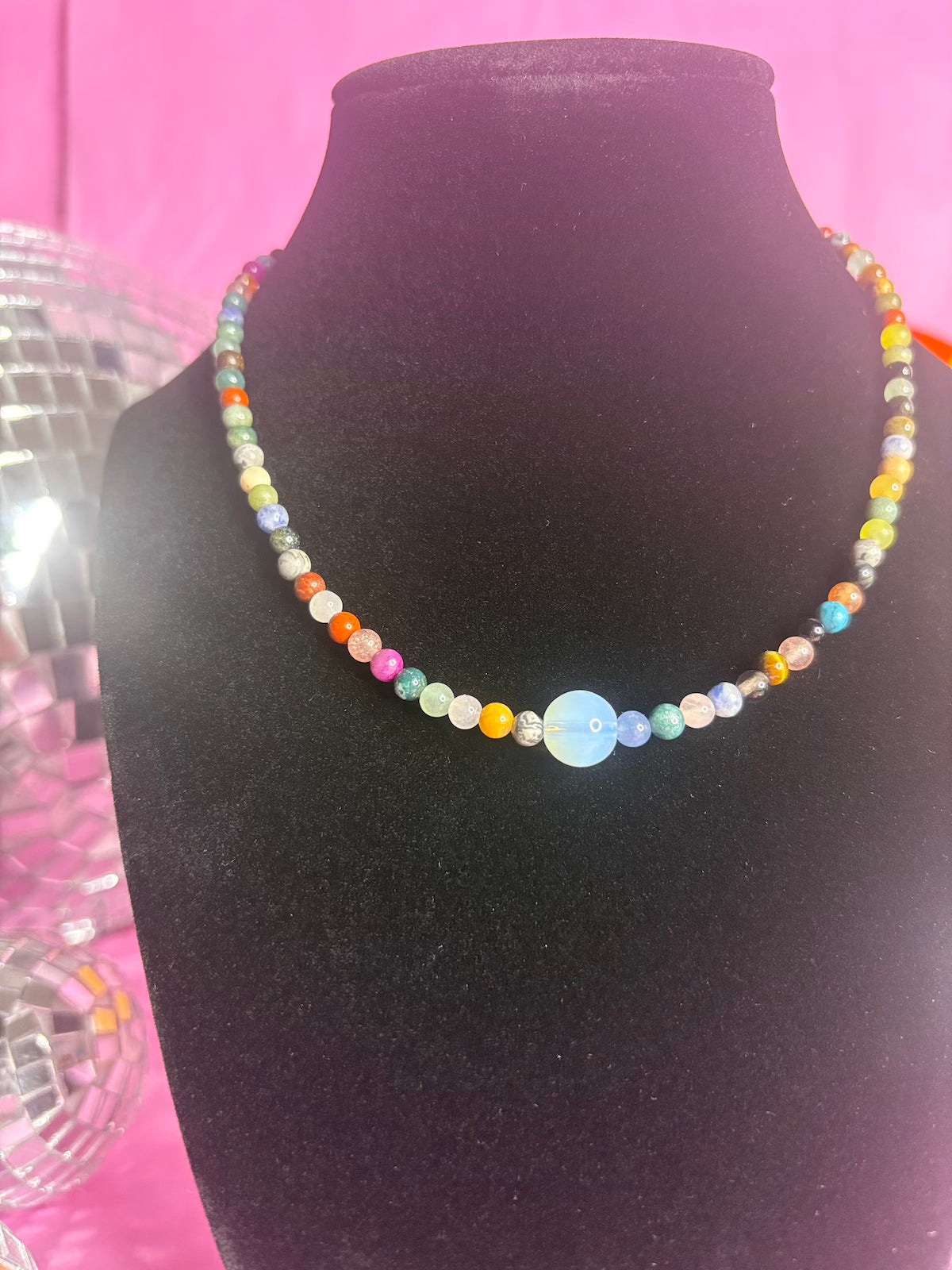 A colorful beaded necklace with a central light blue oval bead, displayed on a black velvet jewelry bust against a bright pink background.