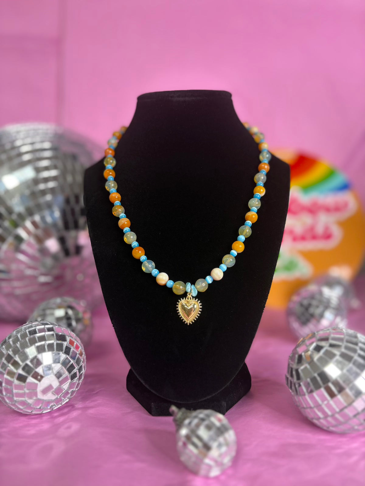 A colorful beaded necklace with a gold heart pendant displayed on a black velvet jewelry bust against a pink background with disco ball props.