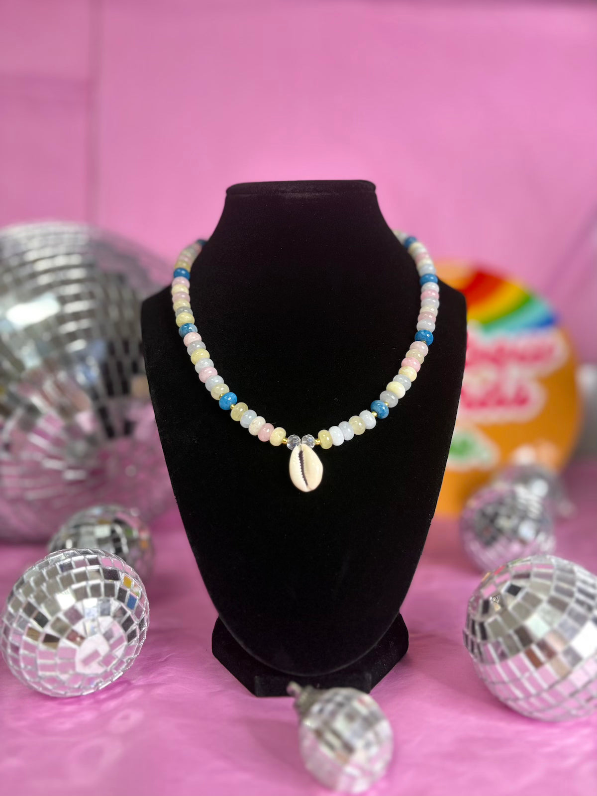 A beaded necklace with a cowrie shell pendant displayed on a black velvet jewelry bust against a pink background, surrounded by disco ball props.
