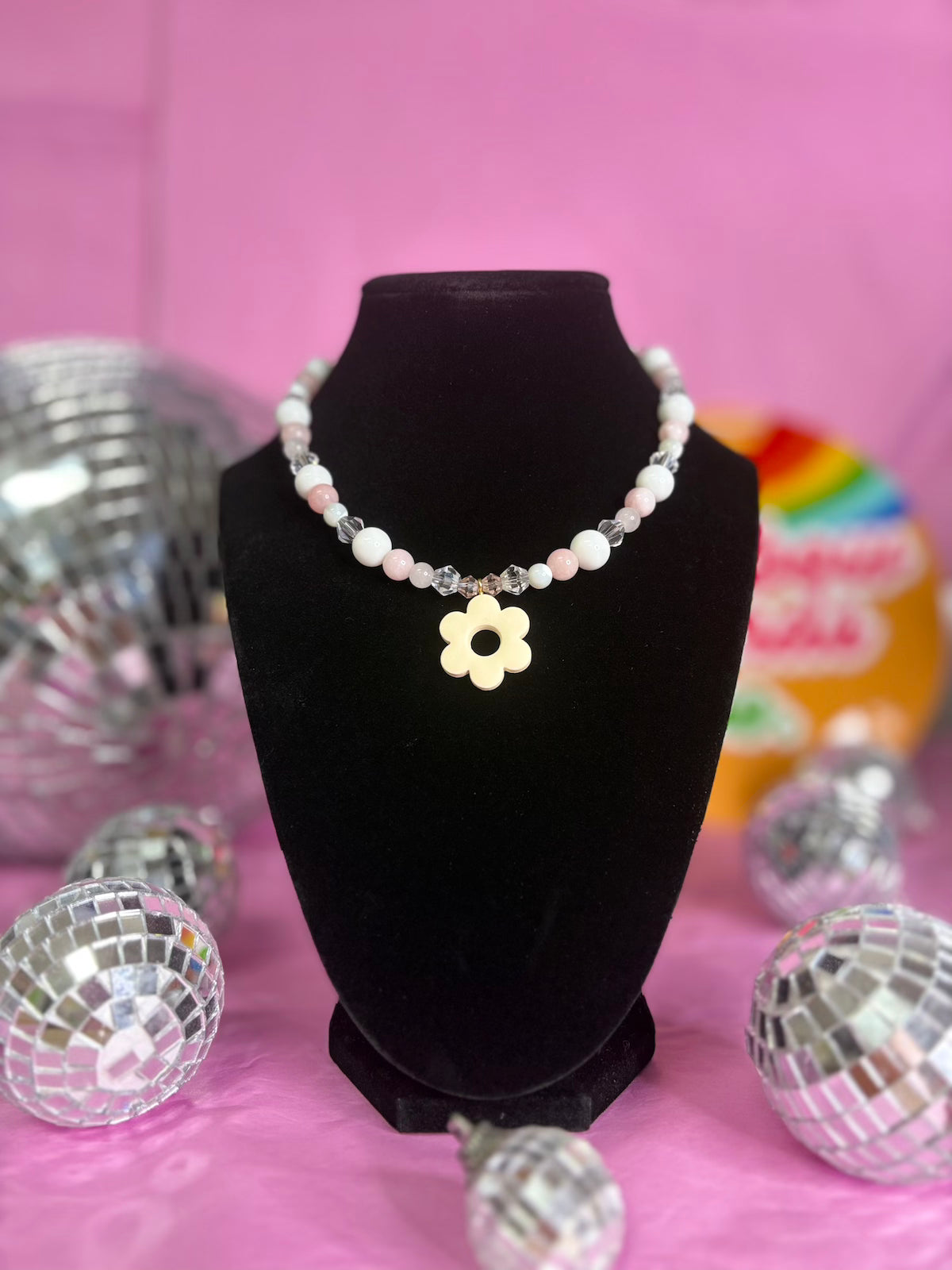A beaded necklace with white and pink pearls, clear rhinestone accents, and a central gold flower charm, displayed on a black velvet bust against a pink background with decorative disco balls.
