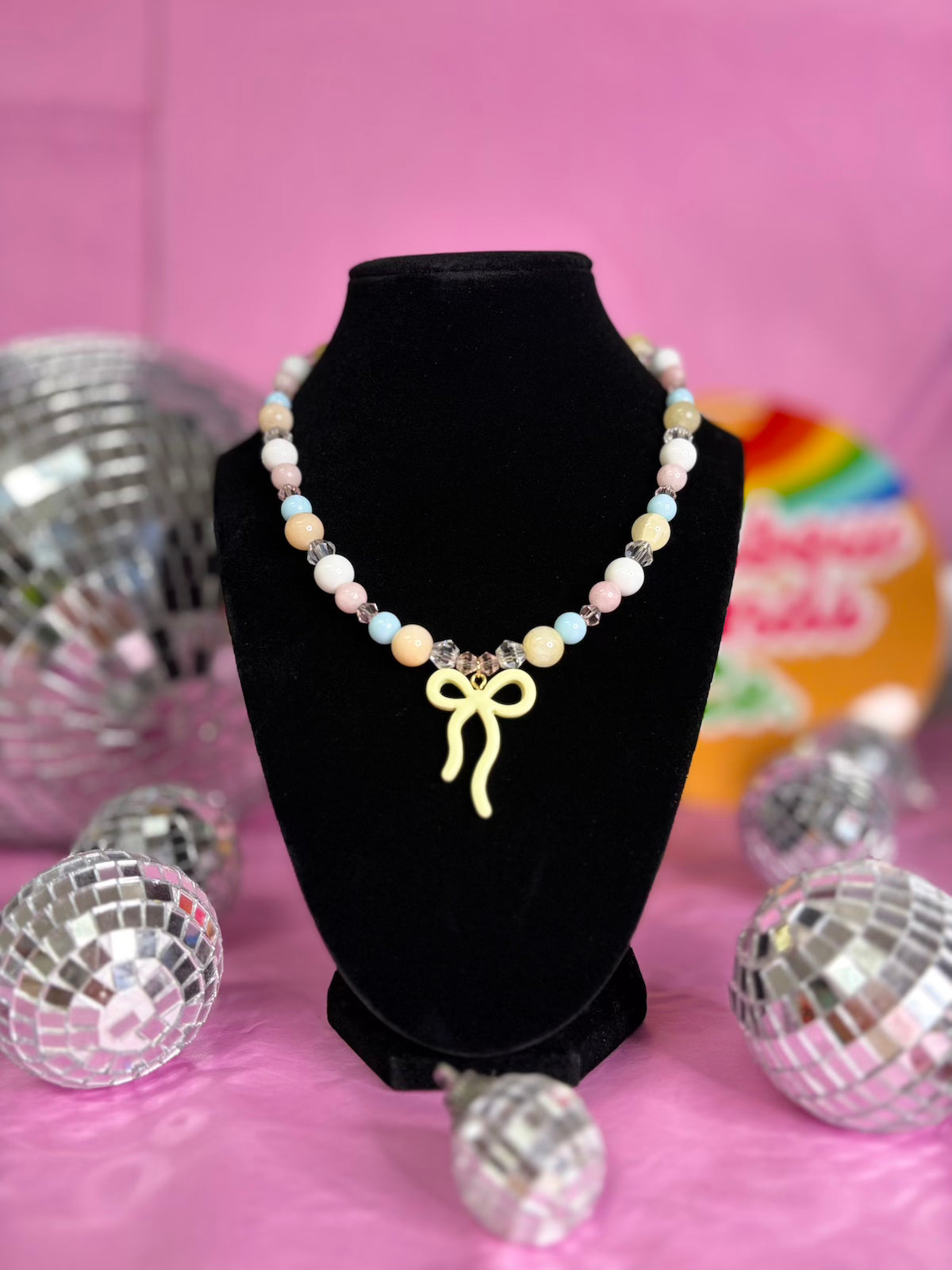 A pastel beaded necklace with a yellow bow charm displayed on a black velvet bust against a pink background with disco ball props.