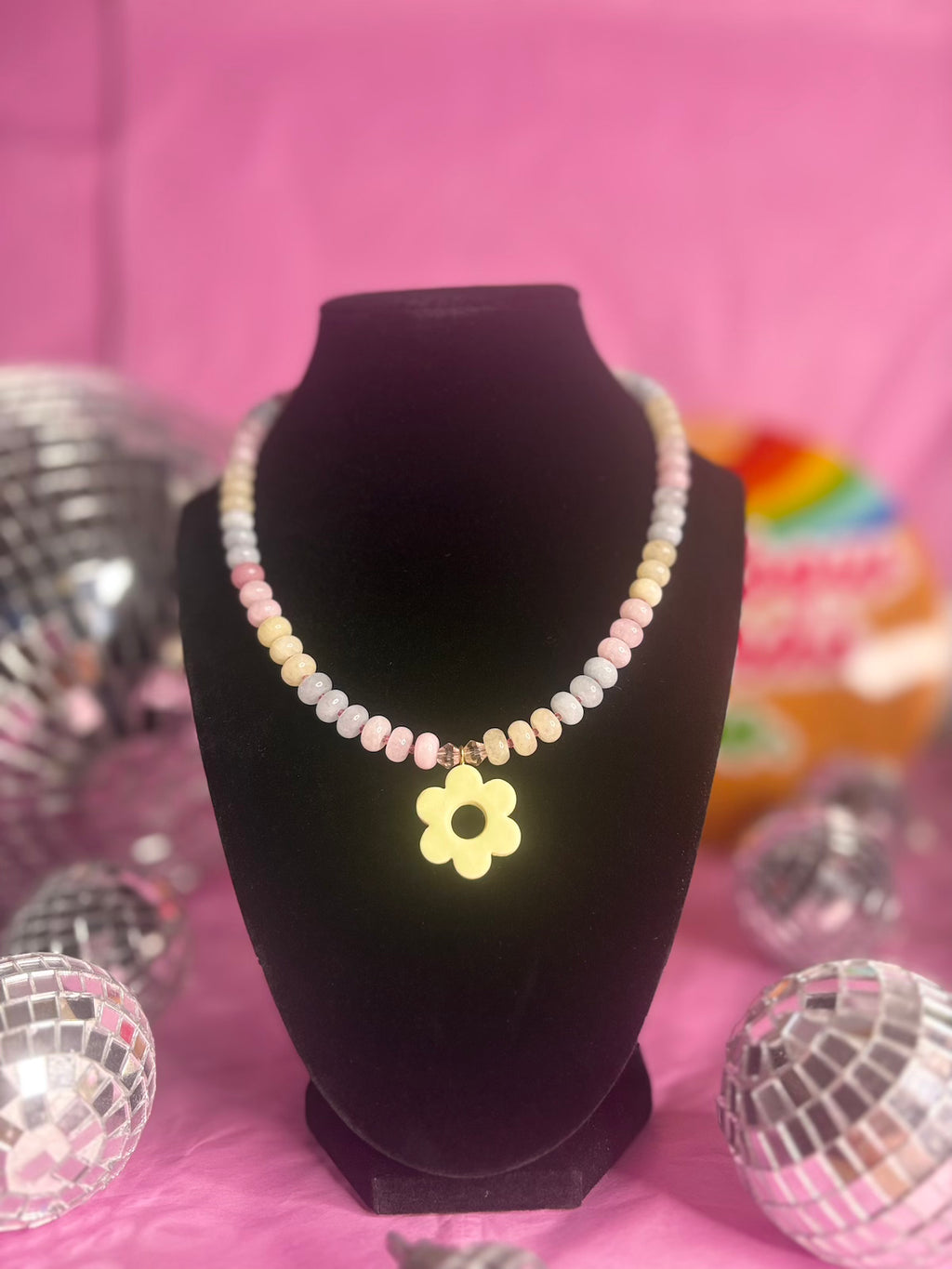 A pastel beaded necklace with a yellow flower-shaped pendant displayed on a black jewelry bust against a pink background, with disco balls as decorative props.