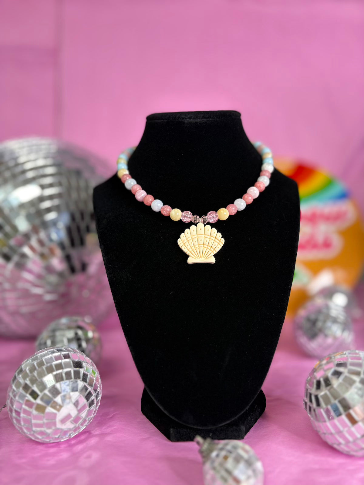 A pastel beaded necklace with a gold seashell pendant displayed on a black velvet jewelry bust against a pink background with decorative disco balls.