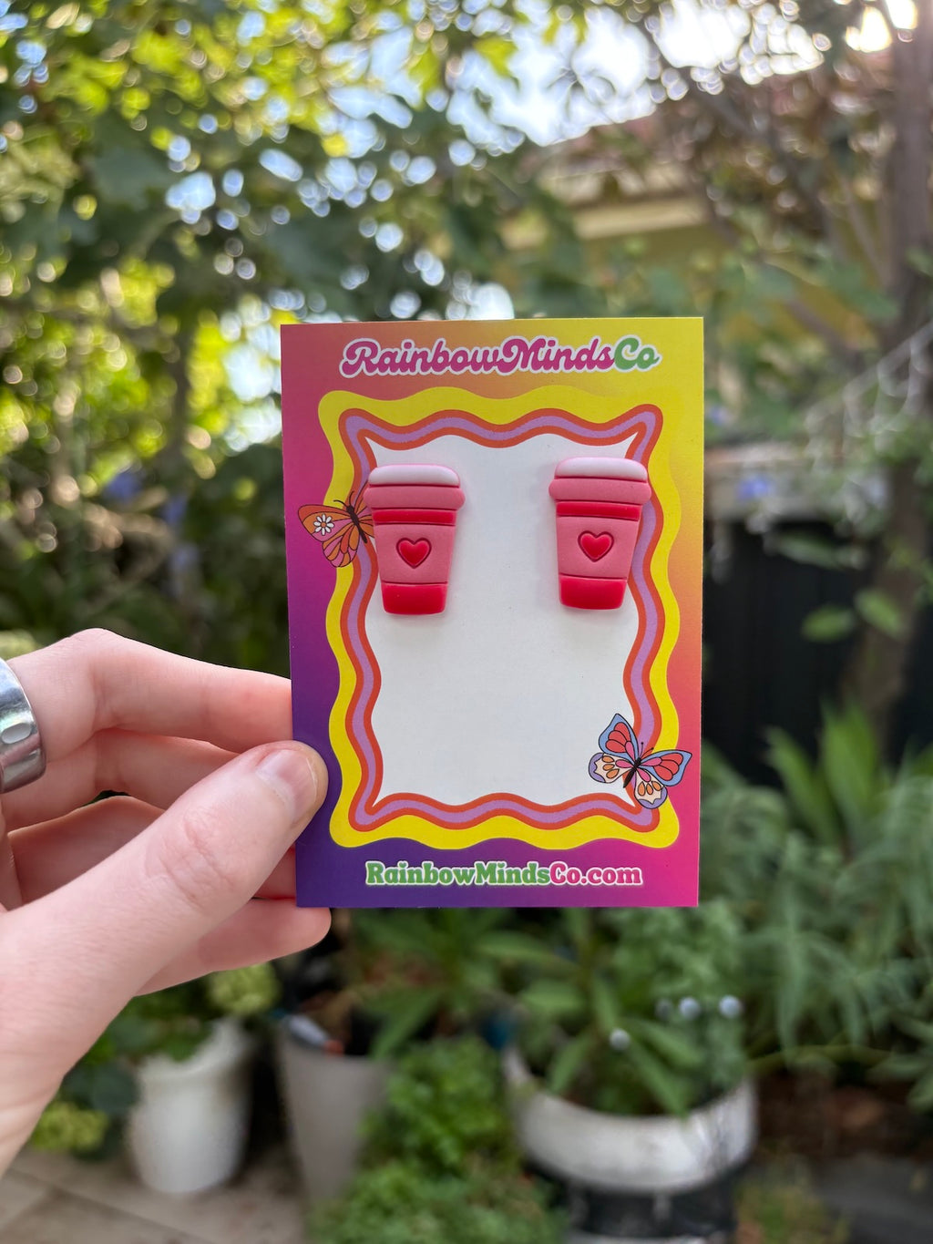 Card with red coffee cup earrings held by a hand against a blurred garden background