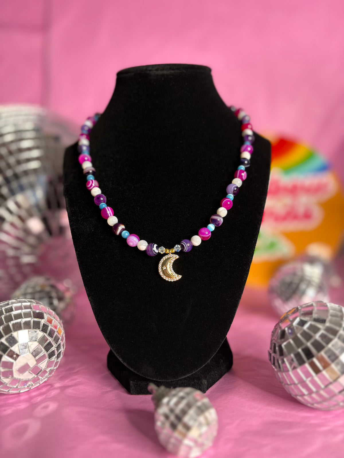 A colorful beaded necklace with a gold crescent moon pendant displayed on a black velvet jewelry bust against a pink background with disco ball props.