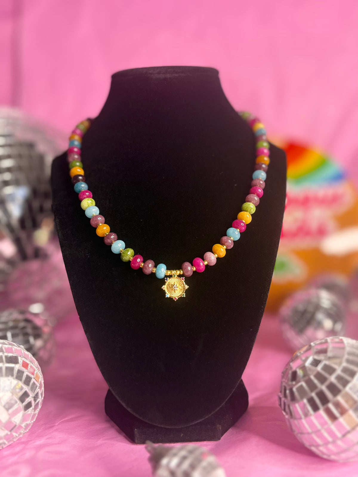 A colorful beaded necklace with a gold sun pendant displayed on a black velvet mannequin bust against a pink background with disco ball props.