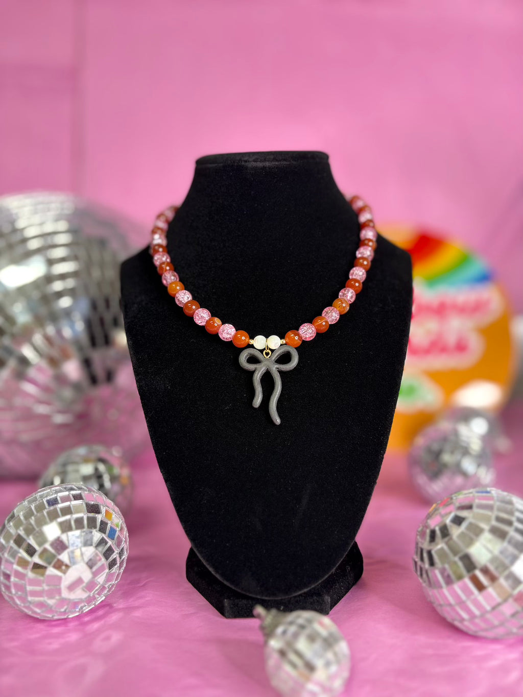 A beaded necklace with pink, orange, and white beads and a silver bow charm, displayed on a black velvet bust against a pink background with disco ball props.