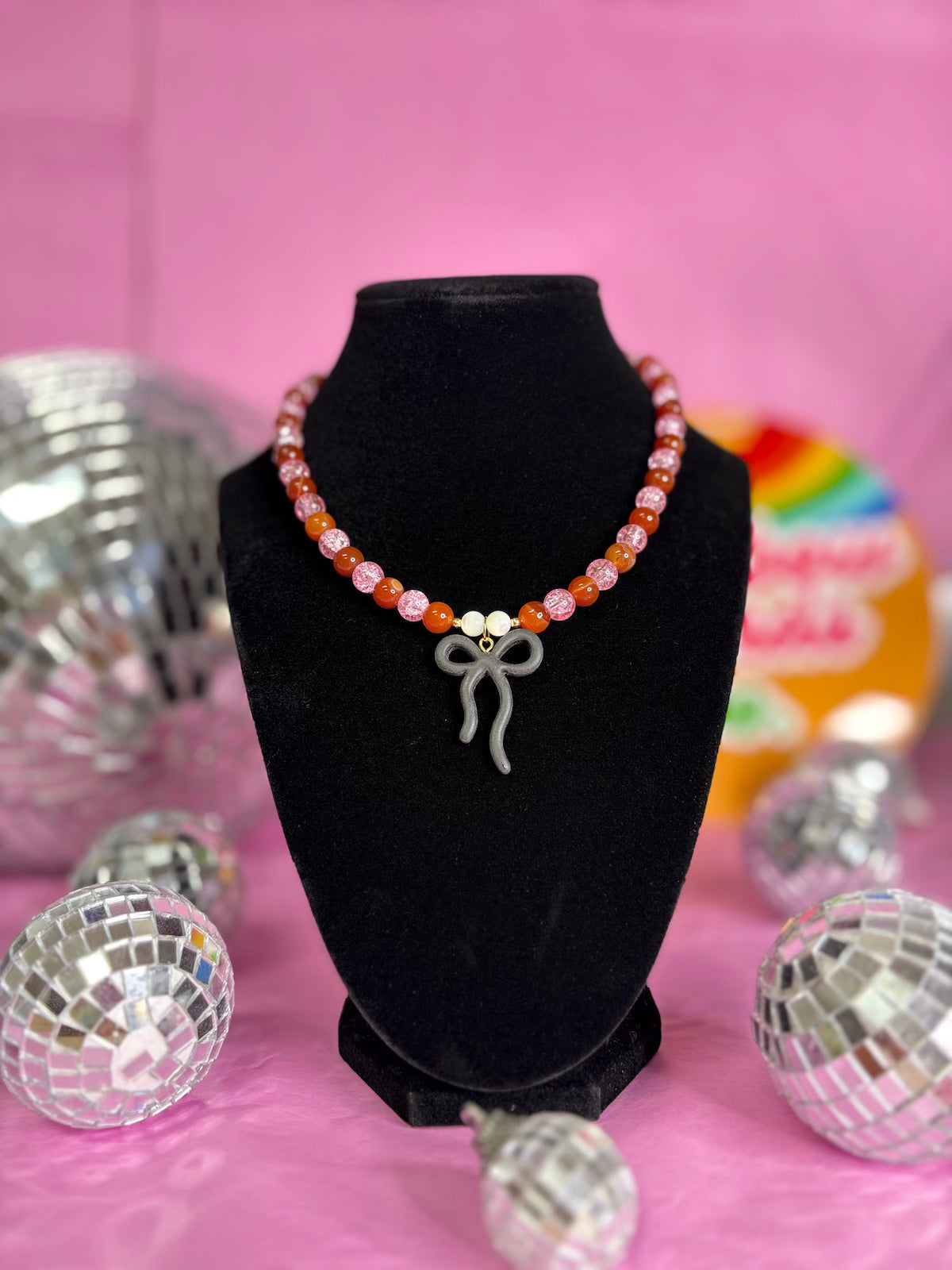 A beaded necklace with pink, orange, and white beads and a silver bow charm, displayed on a black velvet bust against a pink background with disco ball props.