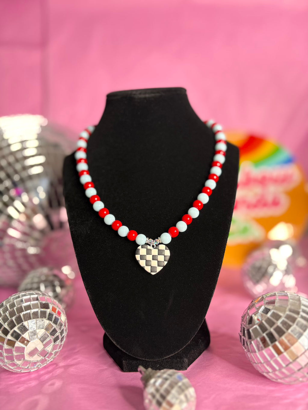 A beaded necklace with red and white beads and a silver heart-shaped pendant featuring a black and white checkerboard pattern, displayed on a black velvet jewelry bust against a pink background with decorative disco balls.